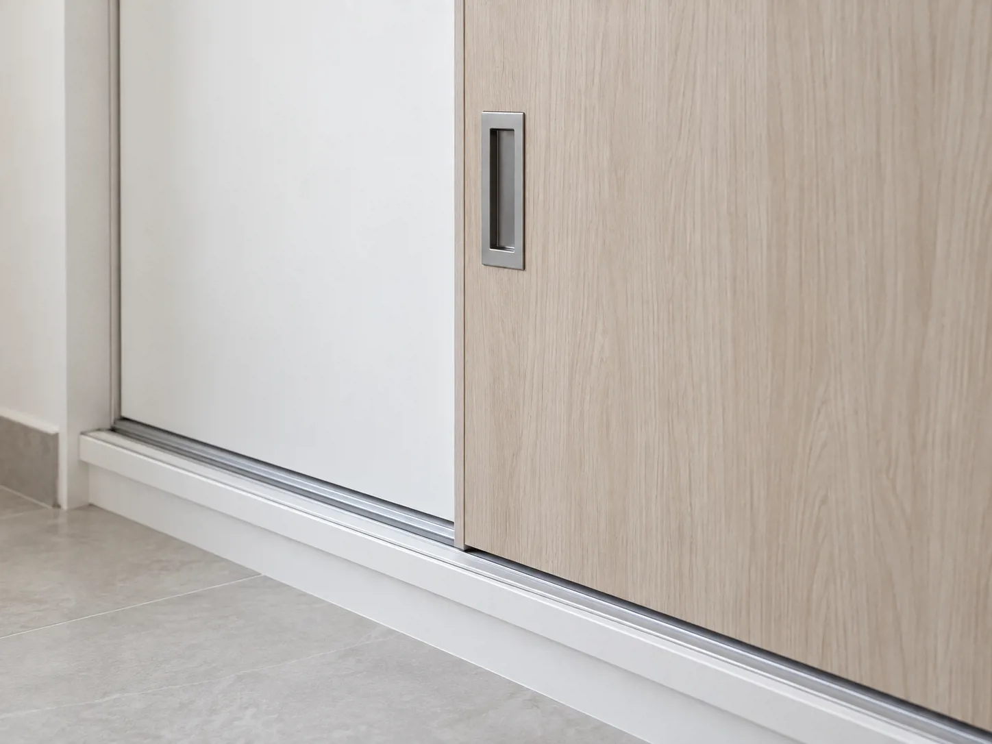 Pintu motif ash dengan tarikan aluminium dan rel bawah menjaga bukaan tetap sejajar dengan muka lemari.